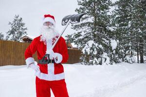 Santa Claus cleans snow with shovel in winter outdoors after a snowfall. Cleaning the streets in the village, clearing the passage for cars, difficult weather conditions for Christmas and New Year photo