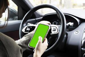Close-up of woman's hands holding smartphone with green empty copy space on screen mockup behind the wheel of sport's car rear view, connecting phone to a electric car. photo