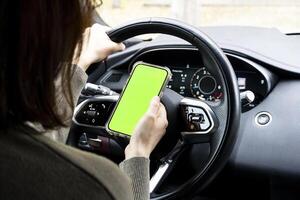Close-up of woman's hands holding smartphone with green empty copy space on screen mockup behind the wheel of sport's car rear view, connecting phone to a electric car. photo