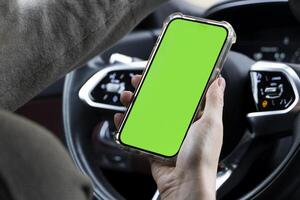 Close-up of woman's hands holding smartphone with green empty copy space on screen mockup behind the wheel of sport's car rear view, connecting phone to a electric car. photo
