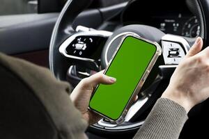 Close-up of woman's hands holding smartphone with green empty copy space on screen mockup behind the wheel of sport's car rear view, connecting phone to a electric car. photo