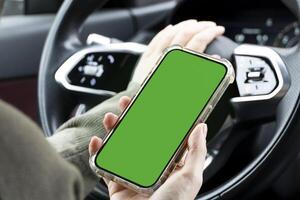 Close-up of woman's hands holding smartphone with green empty copy space on screen mockup behind the wheel of sport's car rear view, connecting phone to a electric car. photo