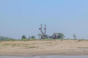 An industrial facility stands prominently on the sandy riverbank, surrounded by sparse greenery and mountains on a sunny day, highlighting the contrast between nature and industry. photo