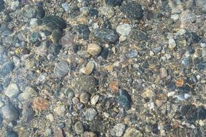 Colorful pebbles spread across the lakebed are visible through the crystal clear water, creating a serene and tranquil atmosphere. The scene captures the calm and beauty of nature. photo