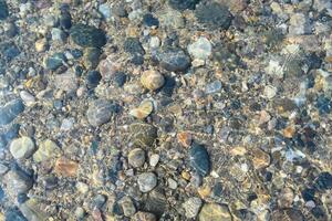 The sunlight shines through clear water, showcasing an array of colorful stones and pebbles on a natural lakeside. This tranquil setting invites relaxation and exploration. photo