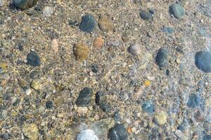 The calm surface of a lake showcases a variety of colorful pebbles submerged beneath clear water, reflecting sunlight on a beautiful day. photo
