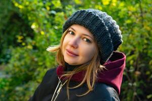 A close-up, side-view portrait of a young woman in a knitted hat and burgundy hoodie, looking thoughtfully into the distance against a vibrant green forest backdrop. Captured with a shallow depth of photo