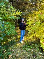 A young woman in a knitted hat and dark jacket stands amidst a lush autumn forest, surrounded by vibrant yellow and green foliage. She holds a small bundle of red leaves, adding a touch of contrast to photo