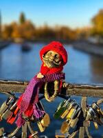 Close-up of a whimsical Homlin figurine dressed in a red hat and scarf, sitting on a bridge railing in Kaliningrad. The background features a blurred river view with vibrant autumn foliage. Captured photo