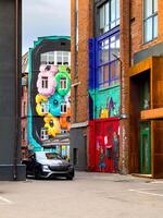A sleek, modern car is parked in an urban alley surrounded by vibrant, large-scale murals on brick walls. The artwork includes bright, abstract designs and lively characters, bringing a pop of color photo