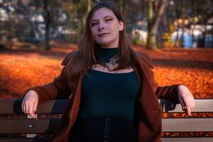 A woman sits confidently on a bench surrounded by autumn leaves in a park during a sunny afternoon photo