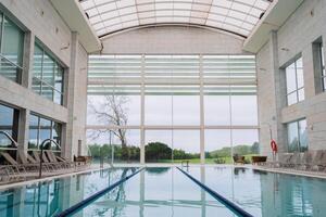 Modern indoor swimming pool with lanes and sun loungers, featuring large glass windows with a view of greenery outside photo