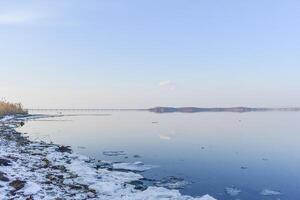 Sea shore covered with melting snow and a mirror-like surface of water reflecting the sky and trees photo