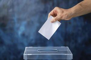 Closeup of a hand casting a white paper ballot into a transparent box on a blue background photo
