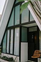 Modern A-frame cabin with large windows and a wicker chair in a cozy outdoor setting. photo