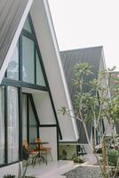 Modern A-frame cabin with large windows and a wicker chair in a cozy outdoor setting. photo