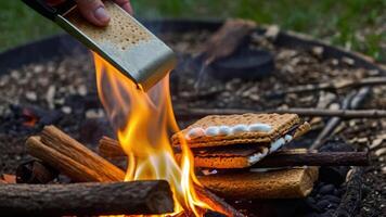 A hand using a stick to toast marshmallows over a campfire for making s'mores. photo