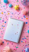Colorful confetti and decorations surrounding a blank spiral notebook on a pink background photo