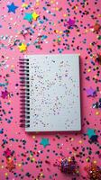 Colorful confetti and decorations surrounding a blank spiral notebook on a pink background photo
