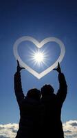 Two people forming a heart shape with their hands against a bright sun in a clear blue sky photo