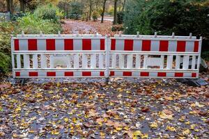 Red and white construction barrier blocking access to park path covered with autumn leaves photo
