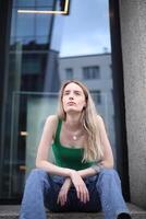 Young Woman in Green Tank Top Sitting Outside a Modern Building in Daylight photo