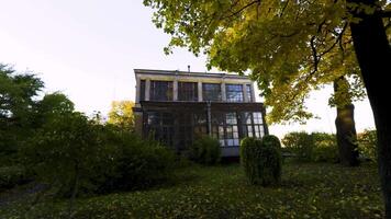 A two-story, weathered house sits peacefully amidst lush greenery and autumn trees. The low-angle shot captures the building partially covered by golden leaves, adding to the tranquil, nostalgic video