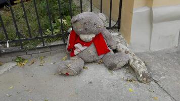 A large, worn teddy bear wearing a red scarf sits abandoned by a metal fence in Saint Petersburg. The bear shows signs of wear, with patches and dirt covering its surface, creating a melancholic video