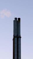 Tall industrial smoke stacks rise against a soft blue sky, with faint wisps of smoke visible. The structure, captured in a minimalistic composition, emphasizes the contrast between the industrial video