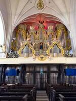 A detailed low-angle view of the historic pipe organ in Konigsberg Cathedral. The baroque design features intricate gold ornamentation and angelic sculptures, framed by soaring gothic arches and photo