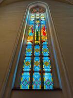A pair of towering stained glass windows inside Konigsberg Cathedral in Kaliningrad, showcasing vibrant religious scenes and intricate geometric designs. The colorful panels depict various figures and photo