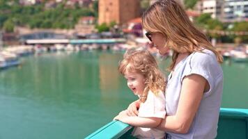 Young mother in sunglasses with small curly blonde daughter goes on cruise on ship leaving sea bay on sunny summer day, they stand on edge of deck and admire panorama, concept of family tripl. video