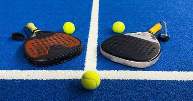 A padel or paddle tennis racket and a bright yellow tennis ball lie on a blue court. The net and a blurred figure of a player are visible in the background. photo