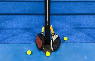 A padel or paddle tennis racket and a bright yellow tennis ball lie on a blue court. The net and a blurred figure of a player are visible in the background. photo