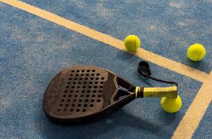 A padel or paddle tennis racket and a bright yellow tennis ball lie on a blue court. The net and a blurred figure of a player are visible in the background. photo