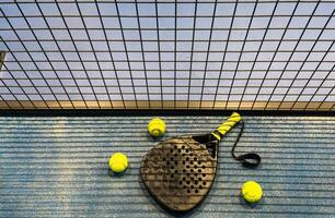 A padel or paddle tennis racket and a bright yellow tennis ball lie on a blue court. The net and a blurred figure of a player are visible in the background. photo