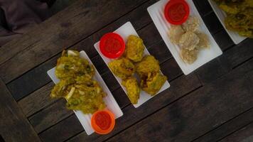 A top-down view of an assortment of Indonesian street food served on a rustic wooden table. A fried banana with grated cheese on top, a plate of vegetable fritters. photo