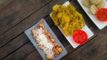 A top-down view of an assortment of Indonesian street food served on a rustic wooden table. A fried banana with grated cheese on top, a plate of vegetable fritters. photo