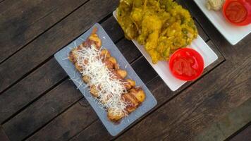 A top-down view of an assortment of Indonesian street food served on a rustic wooden table. A fried banana with grated cheese on top, a plate of vegetable fritters. photo
