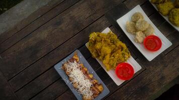 A top-down view of an assortment of Indonesian street food served on a rustic wooden table. A fried banana with grated cheese on top, a plate of vegetable fritters. photo