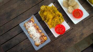 A top-down view of an assortment of Indonesian street food served on a rustic wooden table. A fried banana with grated cheese on top, a plate of vegetable fritters. photo