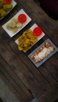 A top-down view of an assortment of Indonesian street food served on a rustic wooden table. A fried banana with grated cheese on top, a plate of vegetable fritters. photo