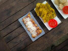 A top-down view of an assortment of Indonesian street food served on a rustic wooden table. A fried banana with grated cheese on top, a plate of vegetable fritters. photo