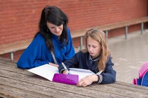 loving mother watching her daughter doing homework on her own at wooden table in children's playground, education concept, back to school, girl in beautiful blue uniform, photo