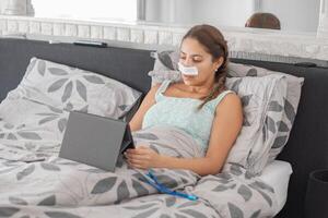Woman resting in bed in casual clothes and bandages on her face after plastic surgery on her nose,using tablet to communicate with family, calm atmosphere and soft colors illustrate recovery process photo
