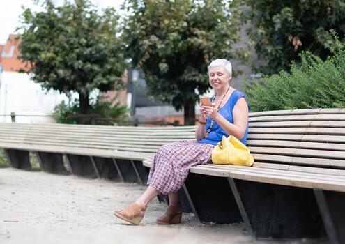 de edad mediana mujer se sienta en un banco en un público sitio en el ciudad y usos un móvil teléfono a comunicar, Jubilación edad, boomers generación, Copiar espacio foto