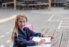 naughty girl making funny faces after successfully completing homework, at wooden table on playground, education concept, back to school, girl in beautiful blue uniform with pink backpack, photo