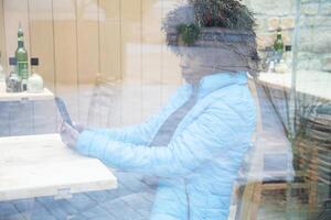 An African American woman in a blue jacket sits behind a window glass in a cafe makes an order using an application on a smartphone, selective soft focus from due to dirty glass. High quality photo