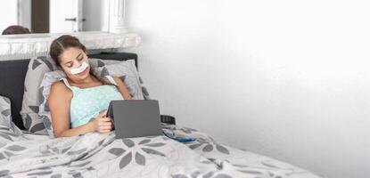 Woman resting in bed in casual clothes and bandages on her face after plastic surgery on her nose,using tablet to communicate with family, calm atmosphere and soft colors illustrate recovery process photo
