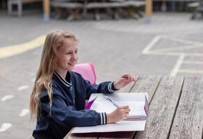 naughty girl making funny faces after successfully completing homework, at wooden table on playground, education concept, back to school, girl in beautiful blue uniform with pink backpack, photo